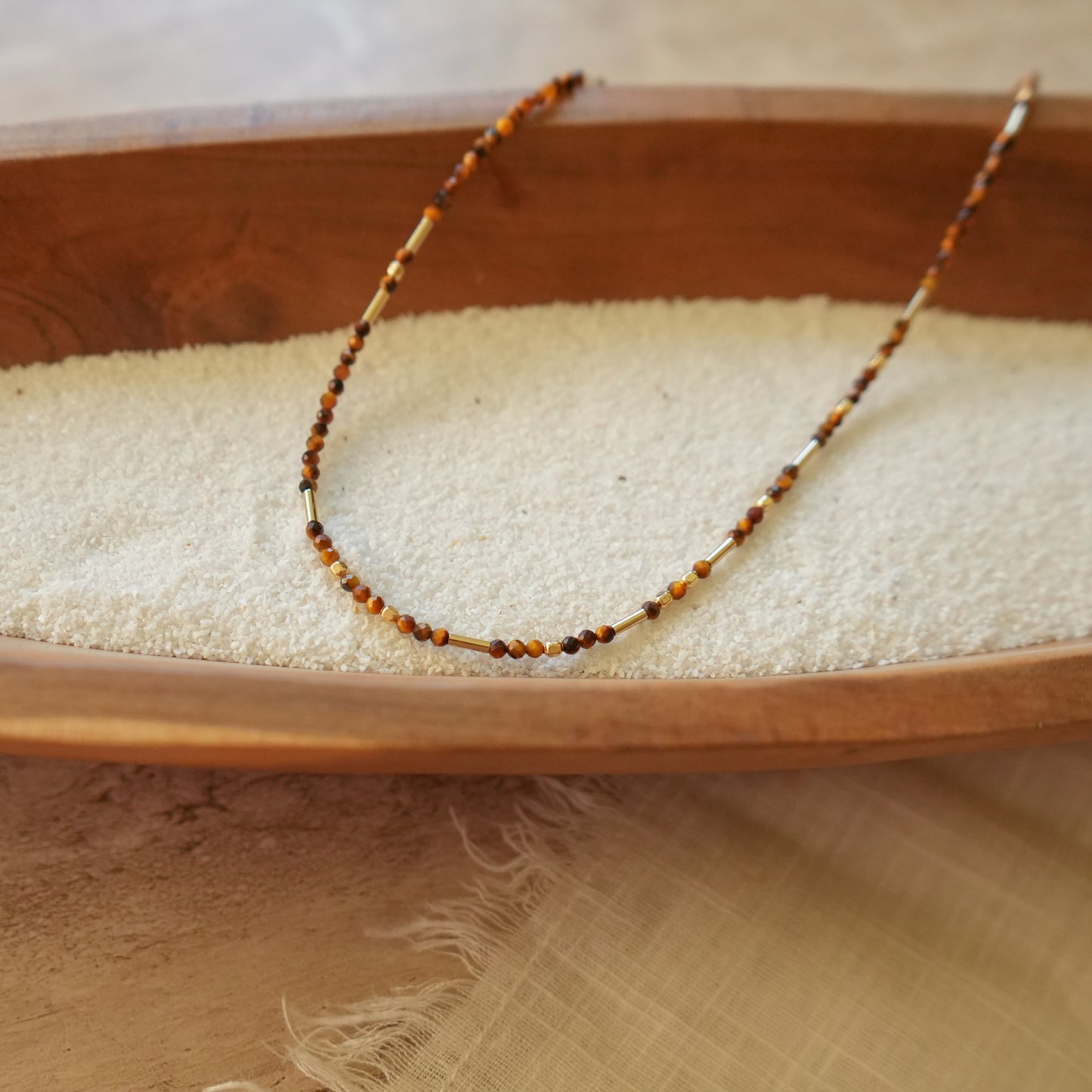 angled view of a tiger's eye and gold-filled beaded necklace on a wooden bowl with white sand