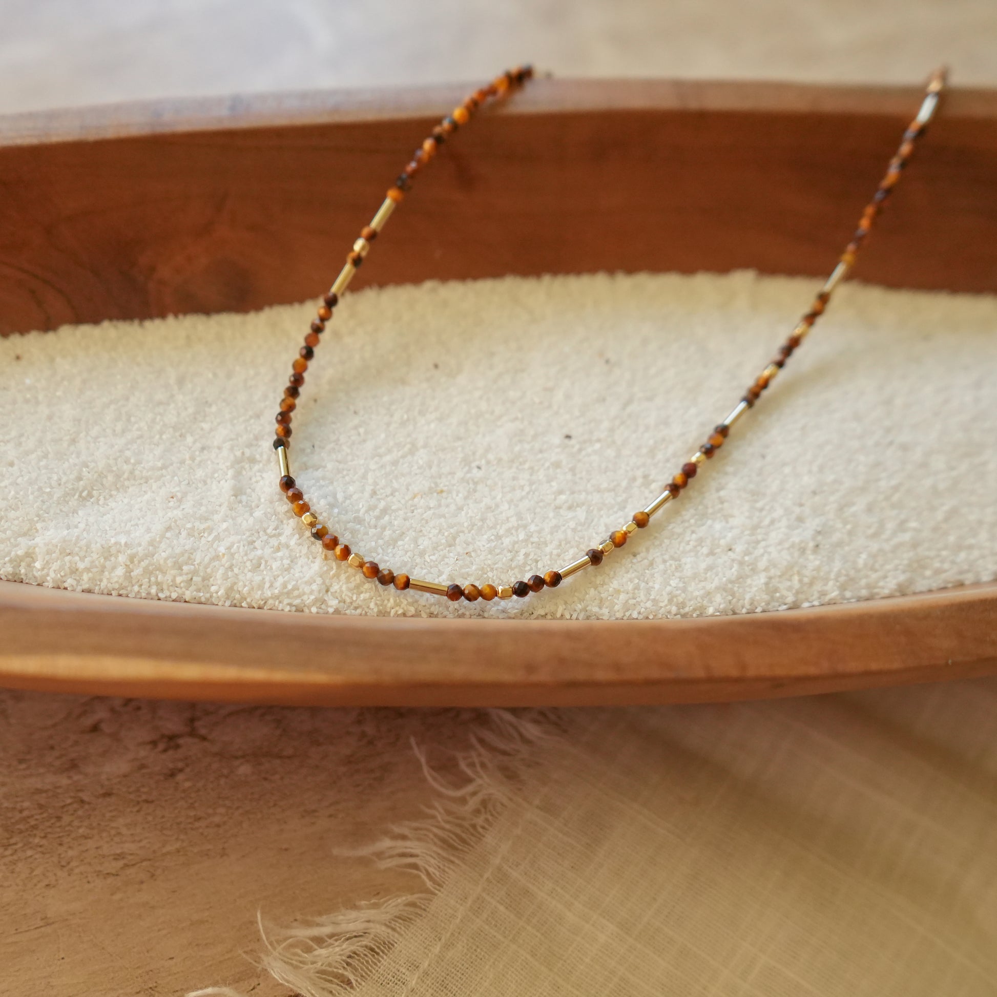 angled view of a tiger's eye and gold-filled beaded necklace on a wooden bowl with white sand