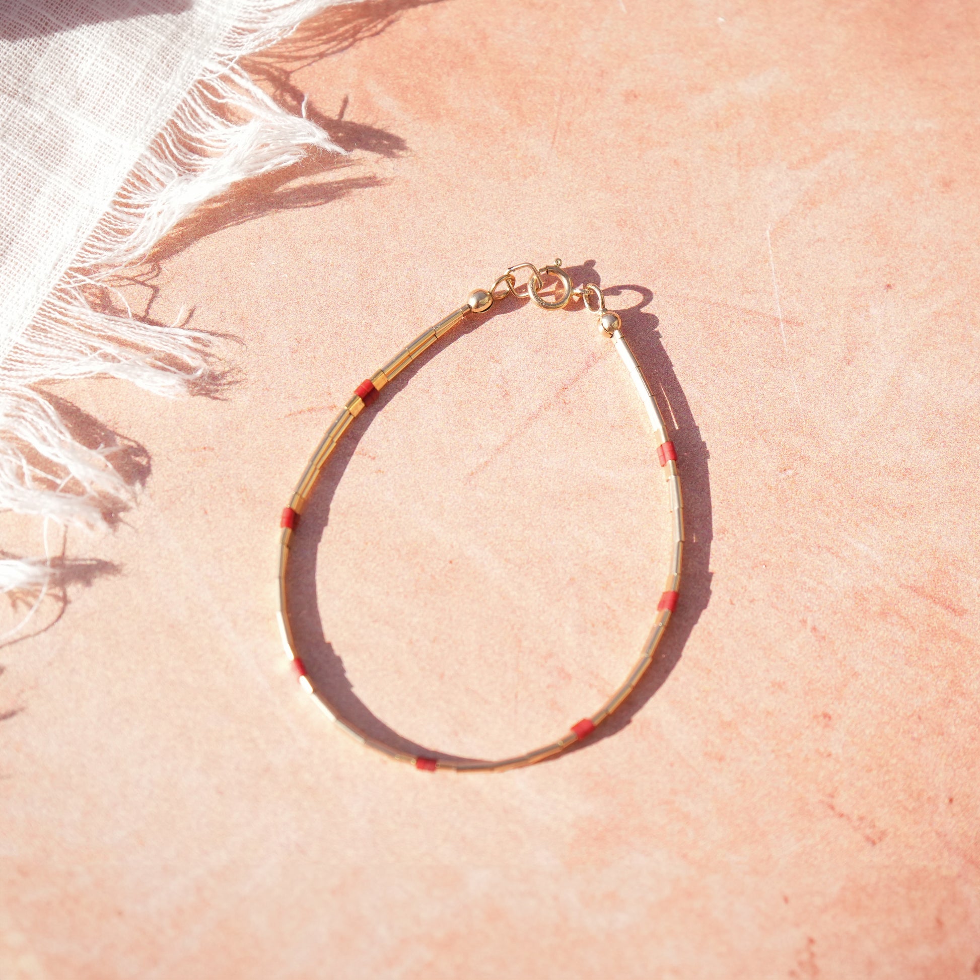 bracelet with 14k gold-fill and red glass beads next to a piece of white cloth resting on a light red surface