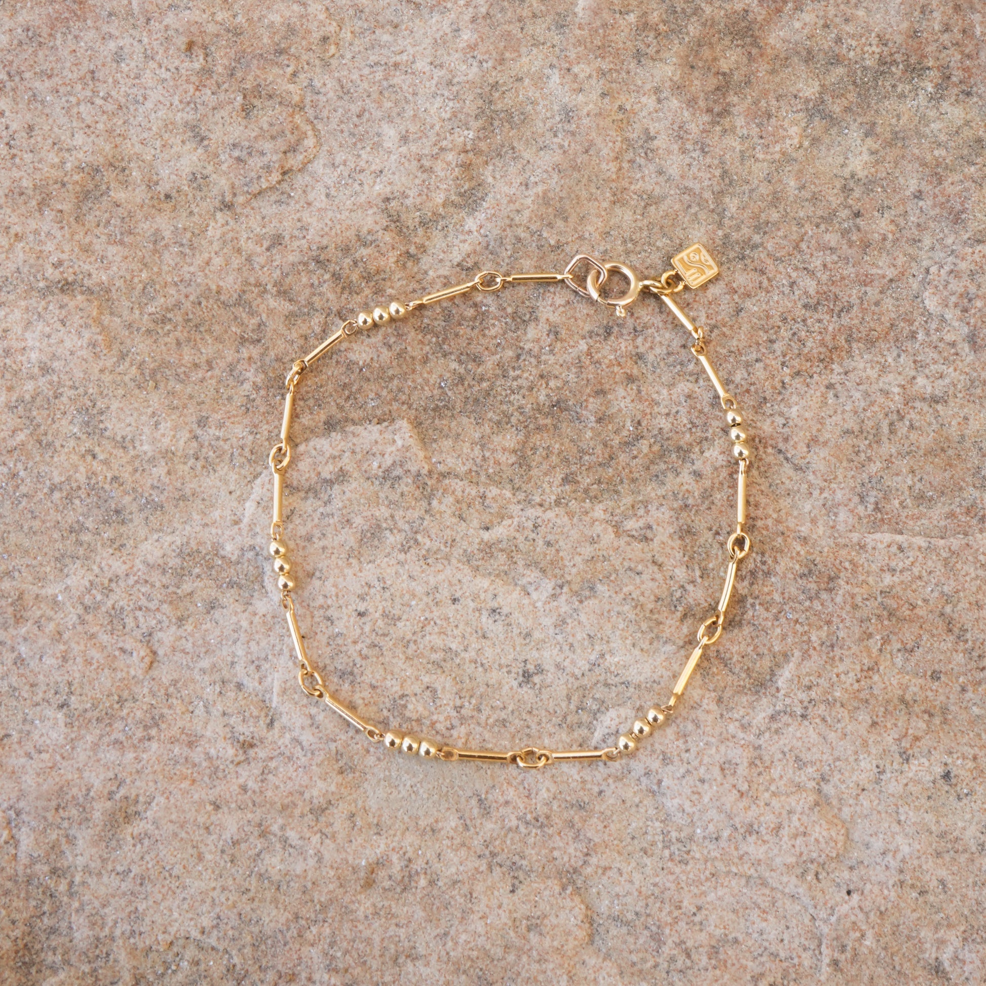 gold-fill beaded bracelet with logo charm on a stone surface